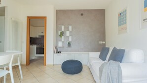 A modern living room with a white sofa, a round ottoman, and a dining area with a white table and chairs. A kitchen is visible in the background.