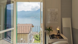 A balcony with a view of a lake and mountains, a framed artwork, a desk with a lamp, and a vase with flowers.