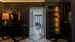 Eine Hotelhalle mit einem Schachbrettmuster aus Marmor, einem Schreibtisch, einer Lampe, einem Spiegel und einer Tür zu einem Balkon mit Blick.