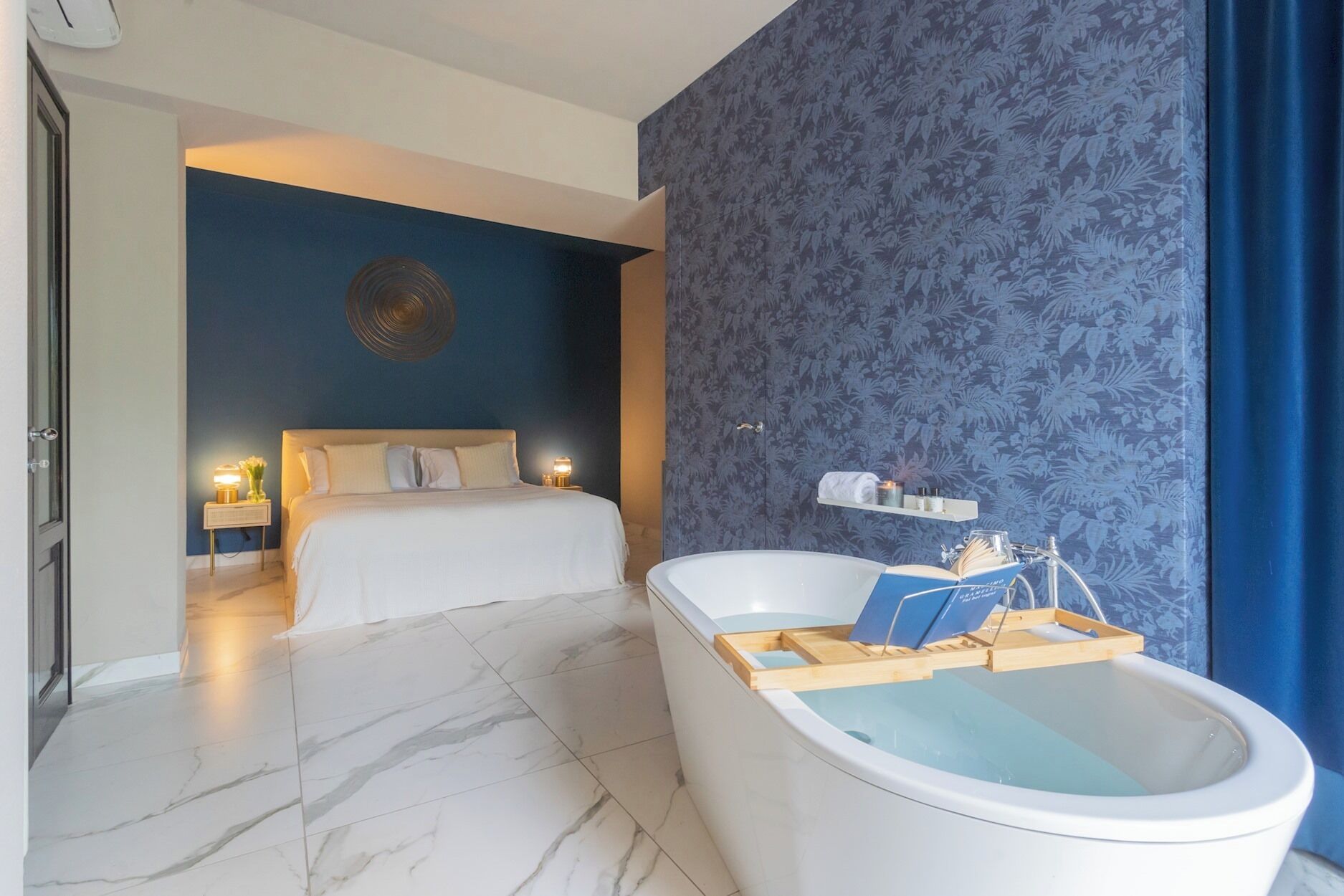 A modern bedroom with a large bed, a bathtub with a wooden tray, and a nightstand with a lamp.