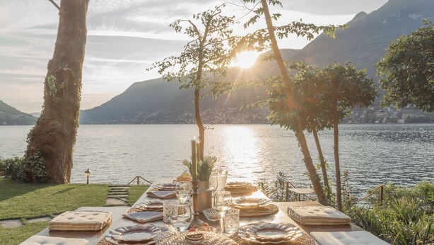 A lakeside dining setup with a table set for two, surrounded by natural scenery and mountains.