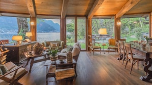 A cozy, wood-paneled room with a large window overlooking a lake and mountains. There are comfortable seating areas, a dining table, and a small desk with a lamp.