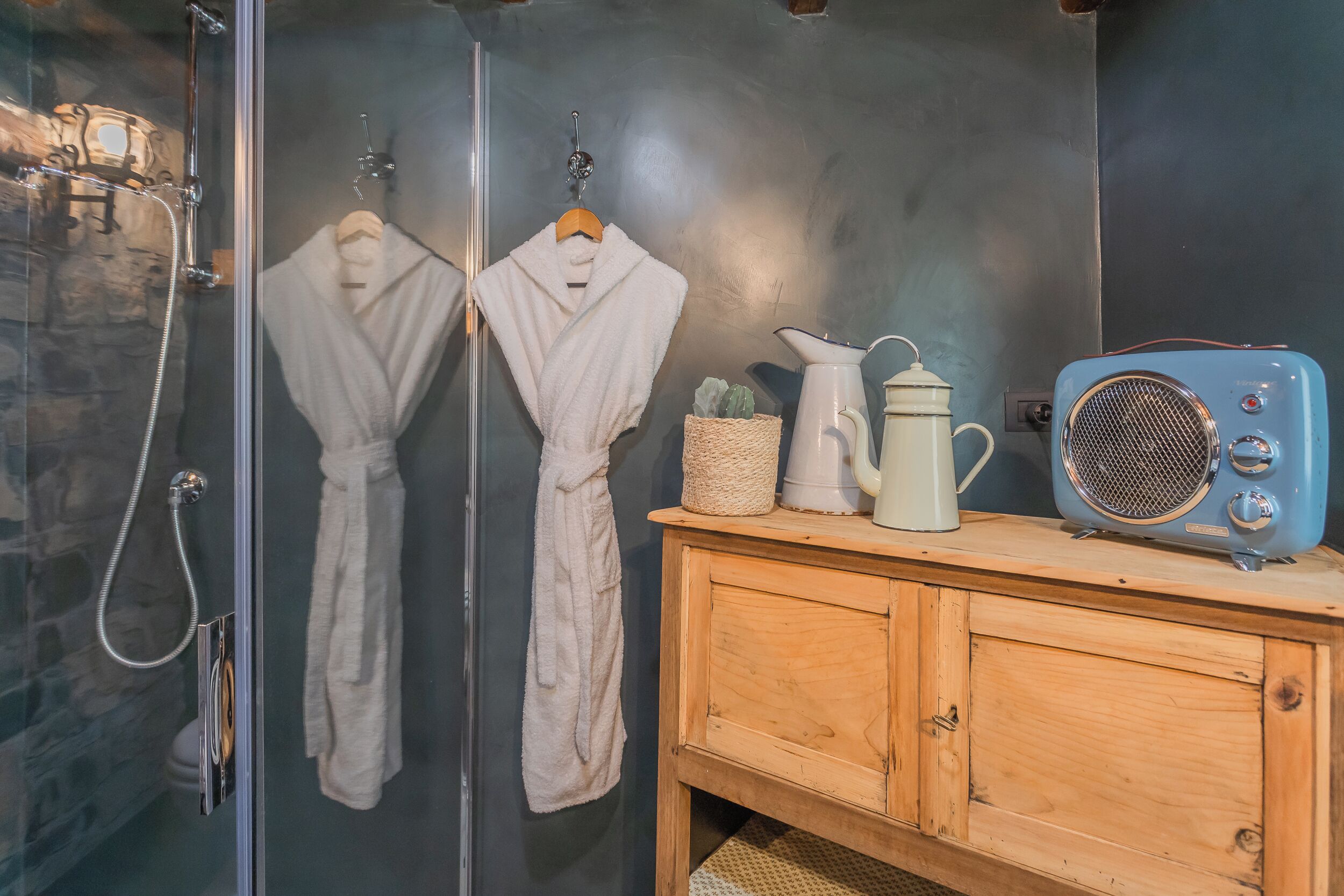 Ein Badezimmer mit Duschkabine aus Glas, zwei weiße Bademäntel an Haken, einem Holzkasten mit einem Vintage-Radio und einem weißen Krug.