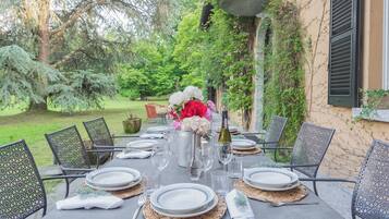 Un área de comedor al aire libre con una mesa preparada para seis personas, rodeada de sillas, en un jardín con árboles y un edificio al fondo.