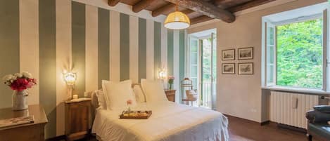 Un dormitorio con cama, mesita de noche, un jarrón con flores, lámpara y ventana con vista a la vegetación.