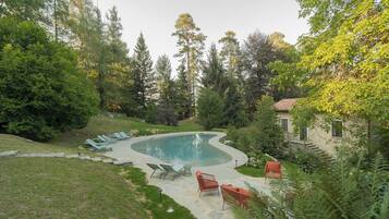 Una piscina con borde curvo, rodeada por un suelo de piedra y césped verde. Hay sillones de descanso y una pequeña silla roja cerca de la piscina.