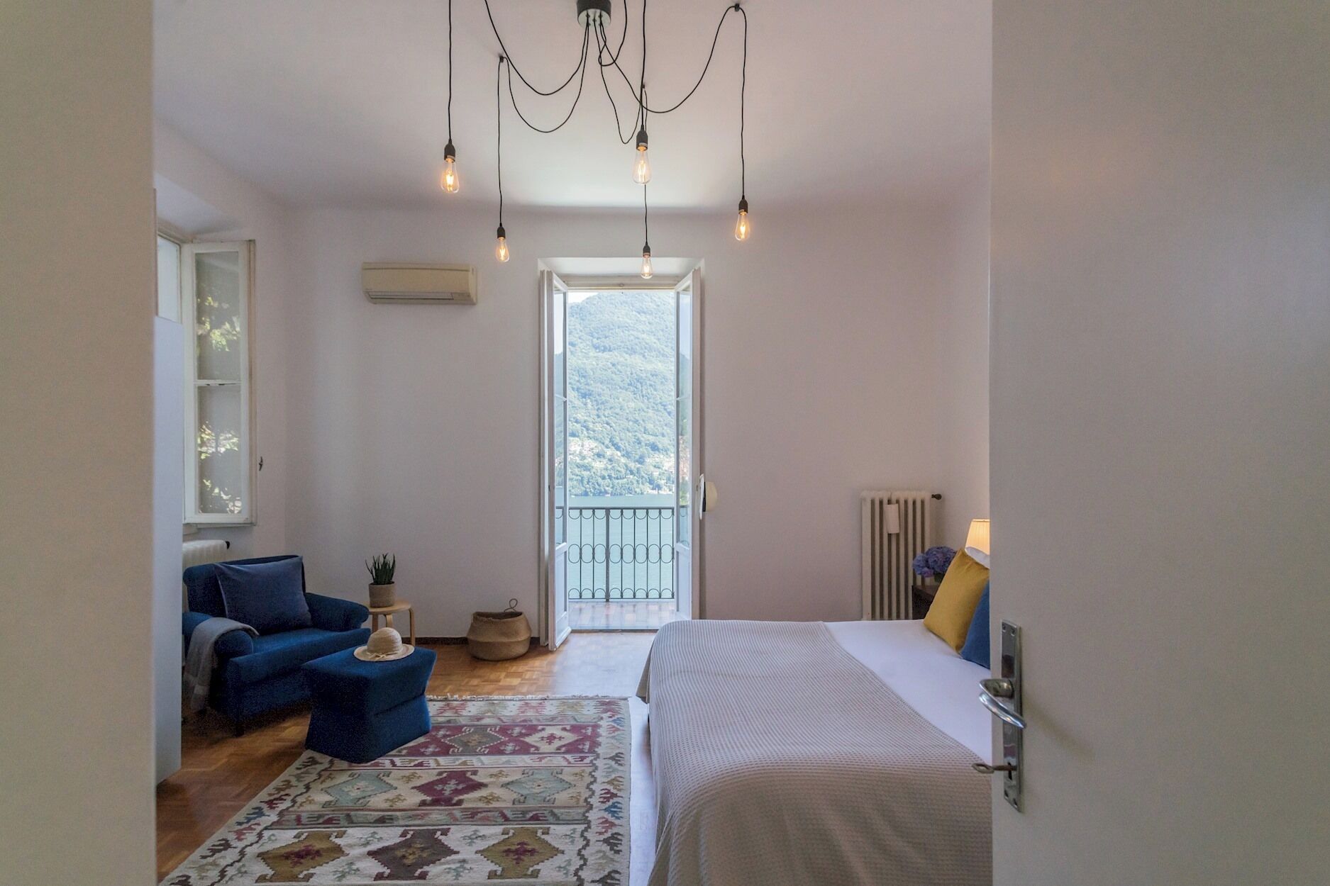 Un dormitorio con cama, sofá azul, una mesita con una planta, alfombra estampada y un balcón con vistas a las montañas.