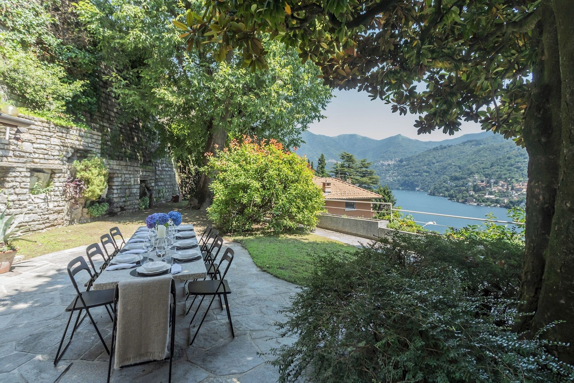 An outdoor dining area with stone flooring, a table set for six with chairs, and a view of a lake and mountains in the background.