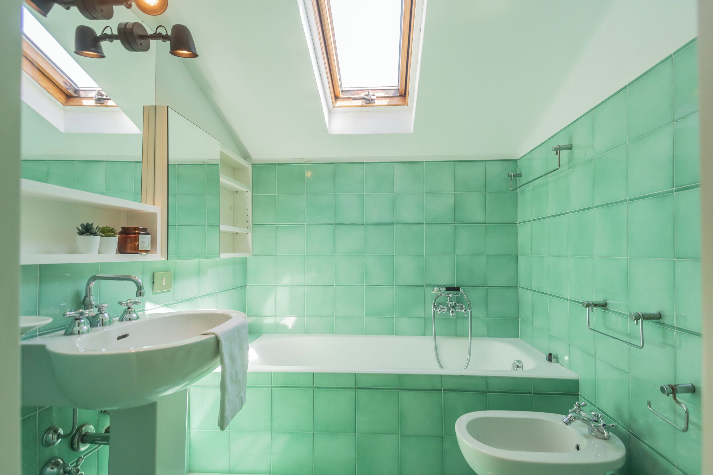 A modern bathroom with green tiles, a skylight, a bathtub, a sink, and a bidet.