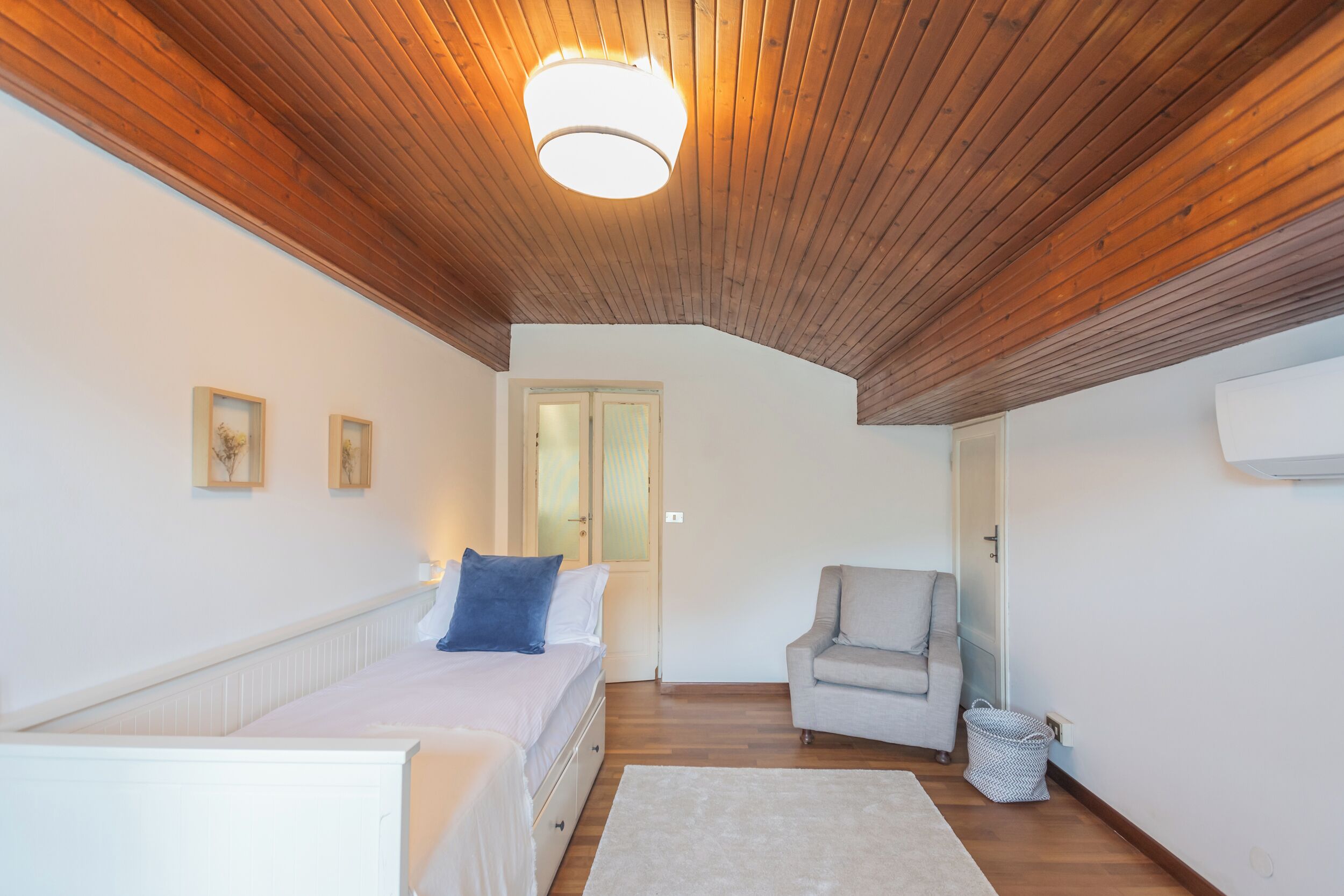 A bedroom with a wooden ceiling, a bed with white bedding and a blue pillow, a grey armchair, a white radiator, and a door leading to another room.