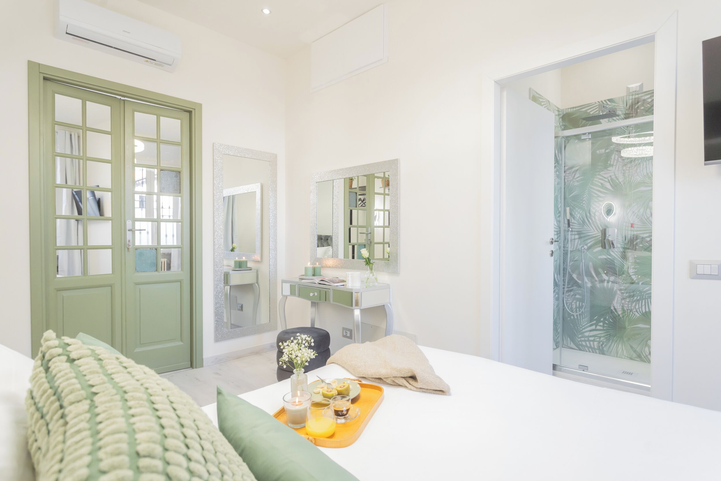 A modern hotel room with a large bed, a vanity with a mirror, a shower with a glass door, and a green door.