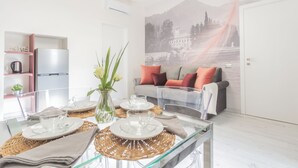 A modern living room with a glass dining table set for four, a sofa with cushions, a kitchen area with a refrigerator, and a mural on the wall. - Cernobbio Dream Suites by Rent all Como (Cernobbio)