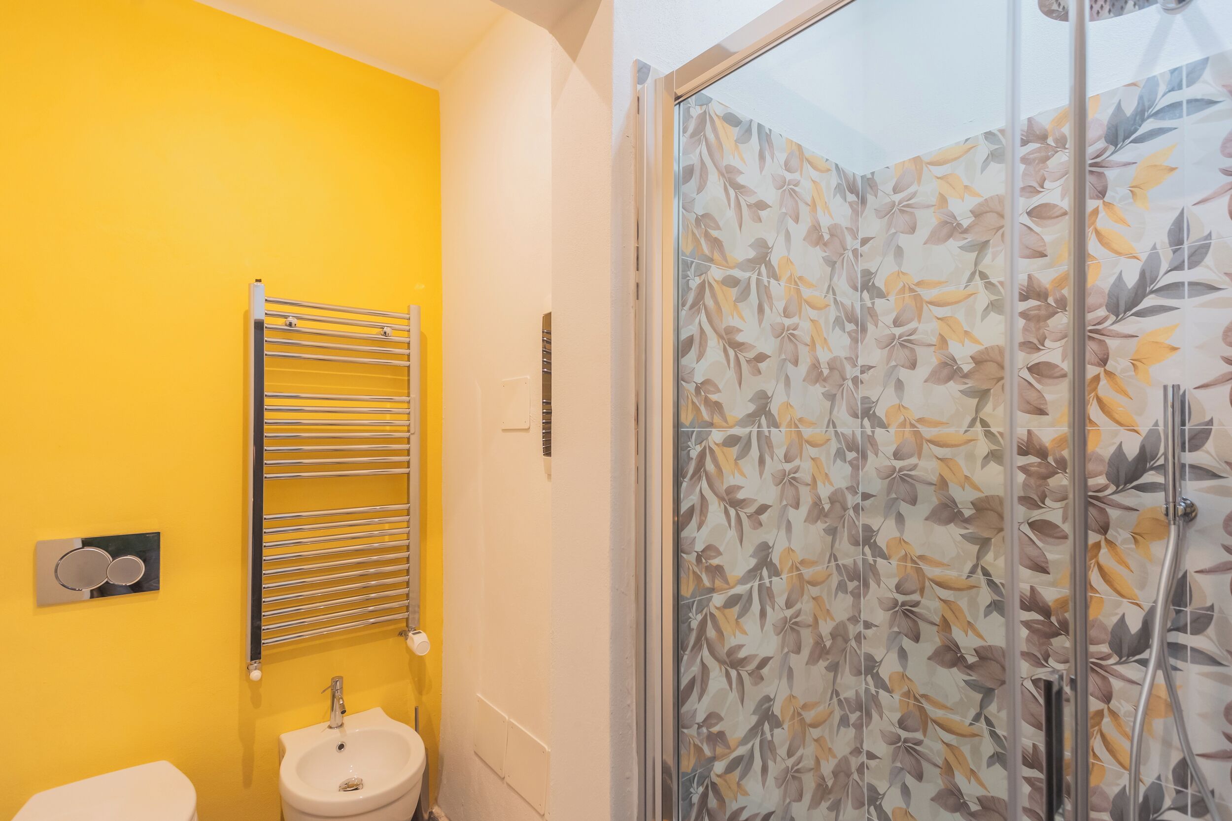 A bathroom with a yellow wall, a white sink, and a glass shower enclosure with leaf-patterned tiles.