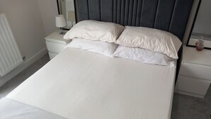 A neatly made bed with white linens and pillows, a bedside table with a lamp, and a nightstand with a vase. - Tranquil Loughton Apartment Retreat (Loughton)