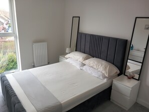 A modern bedroom with a large bed, white bedding, a bedside table with a lamp, a mirror, and a radiator.