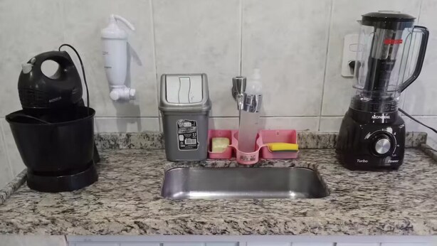 A kitchen with a granite countertop, a sink, a black coffee maker, a white water dispenser, a gray soap dispenser, a pink soap holder, a yellow sponge, and a black blender.