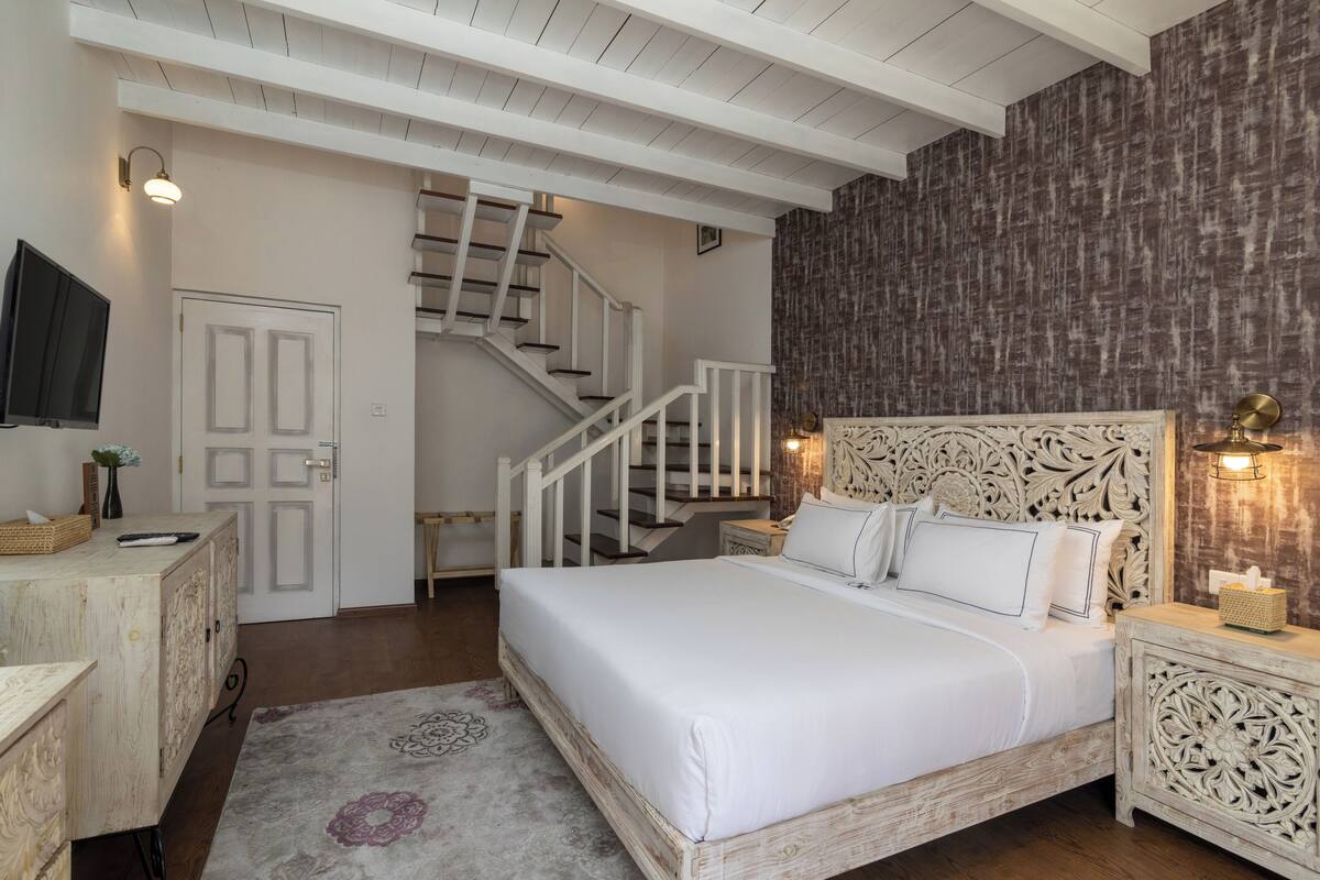 A bedroom with a wooden bed, white bedding, a nightstand with a lamp, a dresser, a TV, and a staircase leading to another room.