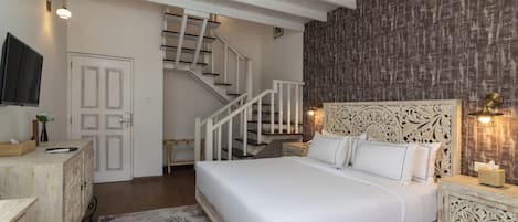 A bedroom with a wooden bed, white bedding, a nightstand with a lamp, a dresser, a TV, and a staircase leading to another room.