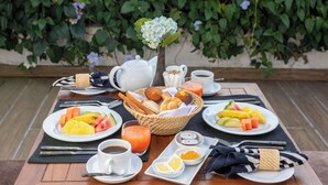 Une table dressée avec des aliments pour le petit-déjeuner, notamment des fruits, du pain et des boissons, avec en arrière-plan une vue sur la verdure et des bâtiments.