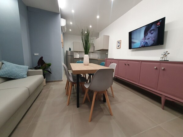 A modern living room with a dining area, a kitchen in the background, and a flat-screen TV mounted on the wall.