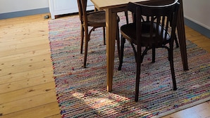 Un comedor con una mesa de madera, cuatro sillas, un tapete colorido, un armario blanco, una tabla de surf y una ventana con vistas.