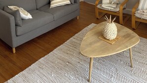 Una sala de estar moderna con un sofá gris, dos sillones de madera, una mesa redonda de centro, un tapiz de pared tejido y una lámpara colgante.