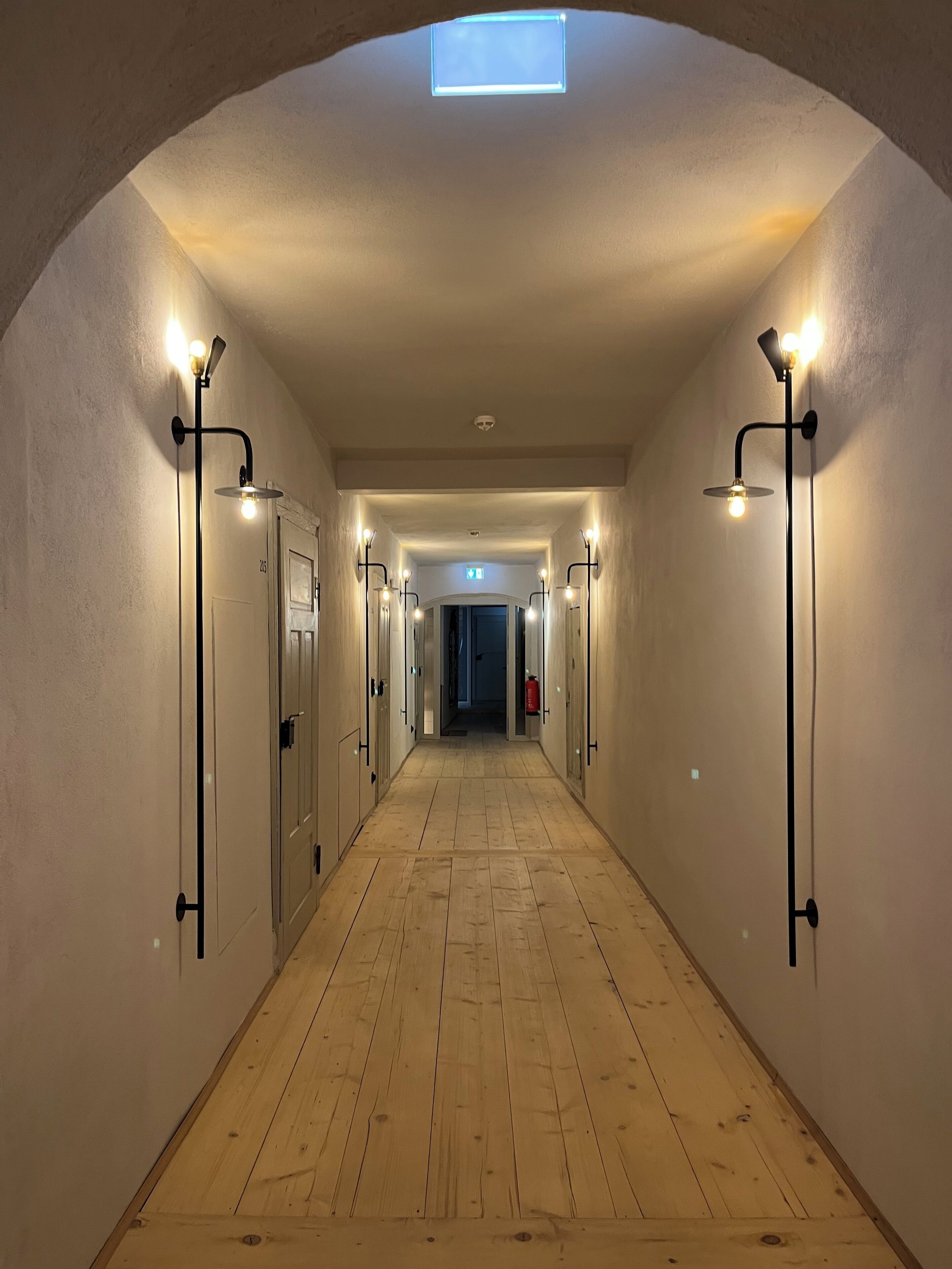 A hallway with wooden flooring, white walls, and multiple light fixtures along the sides.