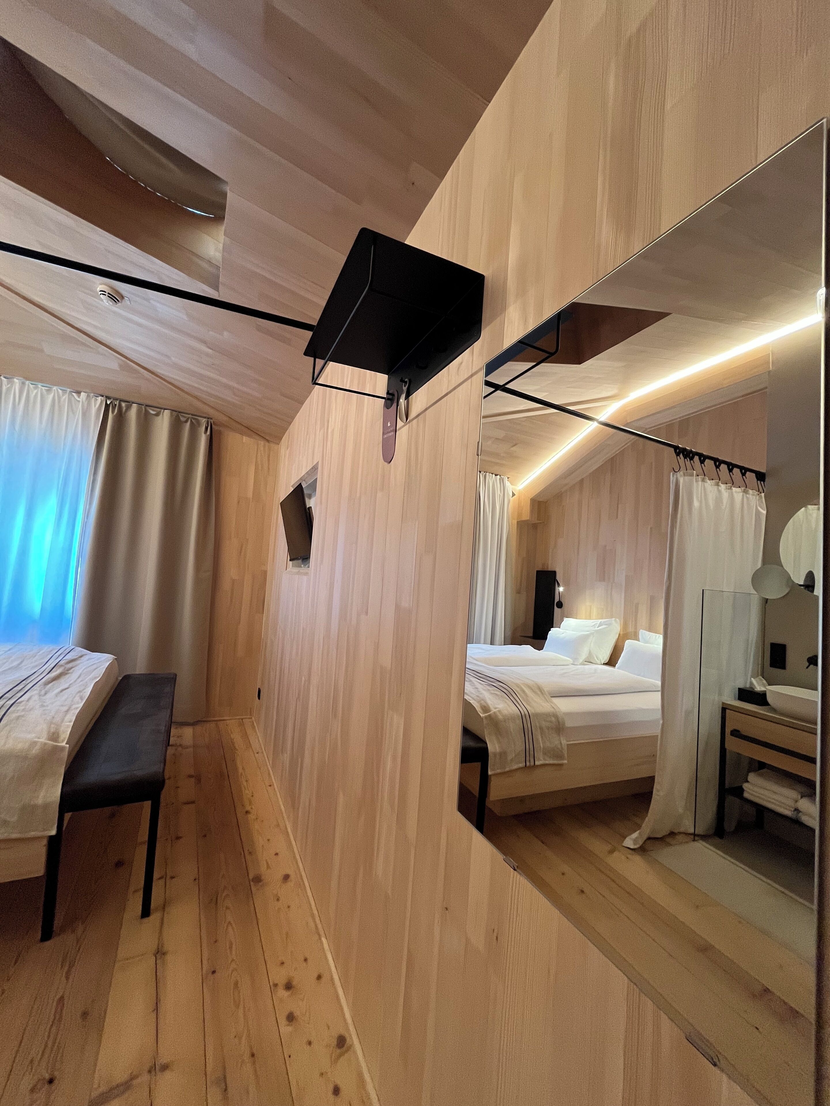 Ein modernes Hotelzimmer mit Holzwandverkleidung, einem großen Spiegel, einem Bett mit weißen Laken, einer Bank und einem kleinen Nachttisch mit Lampe.
