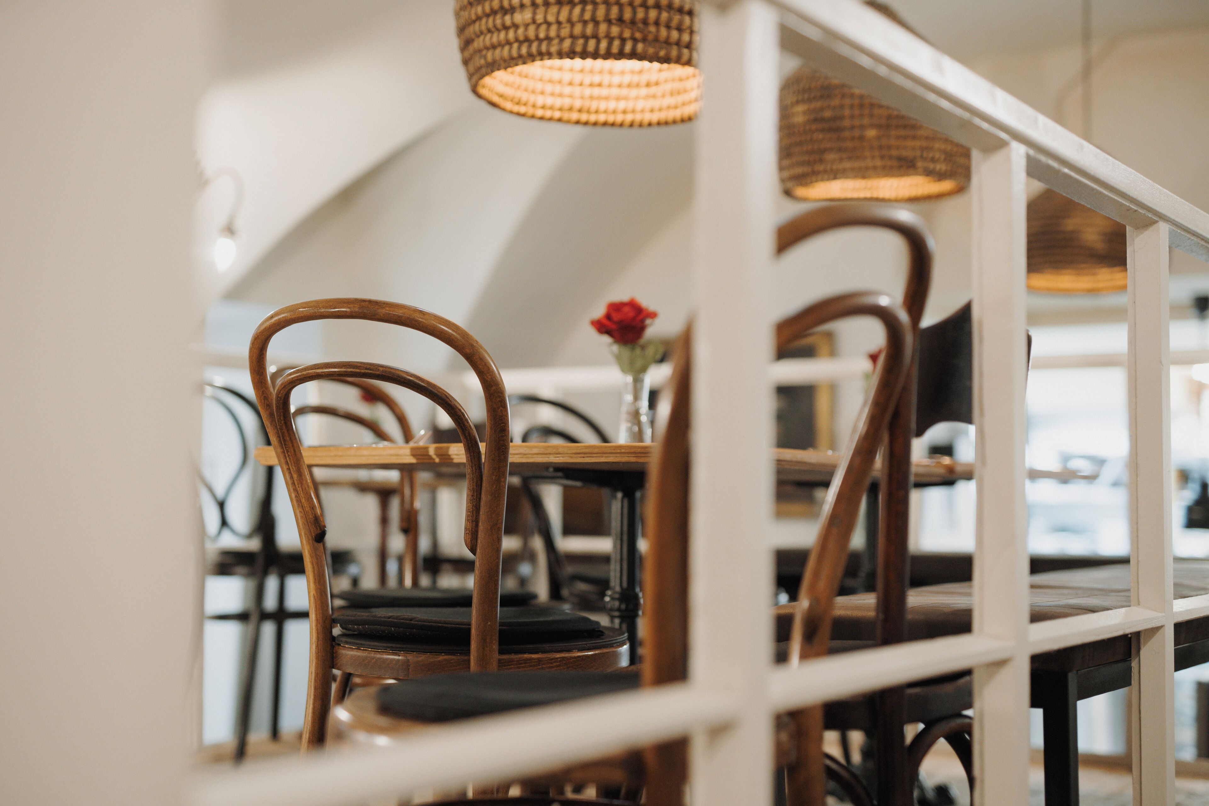 Een eetgedeelte met houten stoelen en tafels, een rieten hanglamp en een vaas met een rode roos.