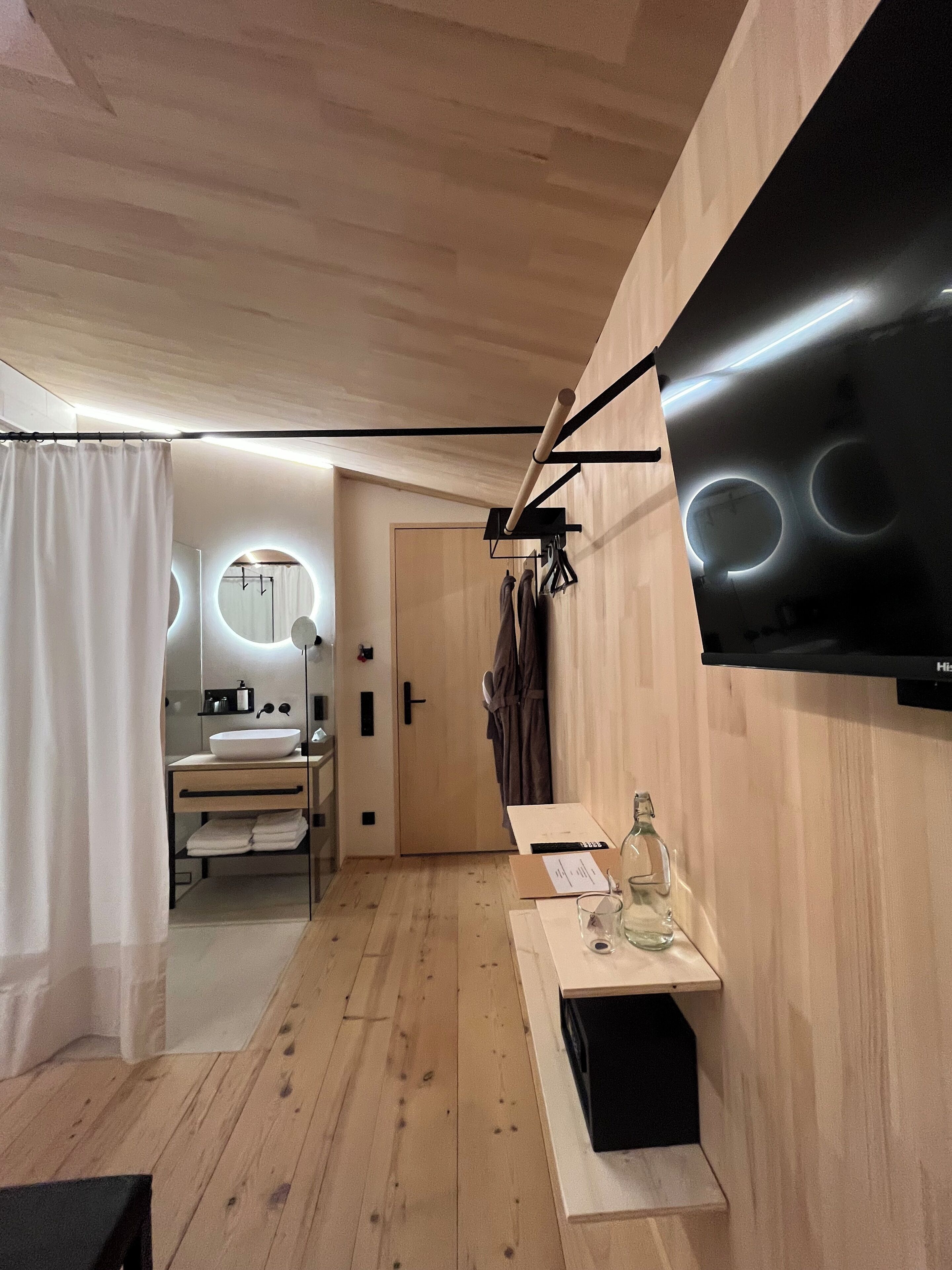 Une salle de bain moderne avec un grand miroir, un lavabo et un rideau de douche. Le sol est en bois et un téléviseur à écran plat est fixé au mur.