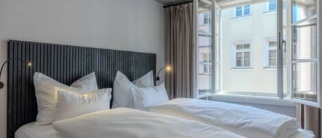 A hotel room with two beds, a wooden headboard, a large window with curtains, and a view of buildings outside.