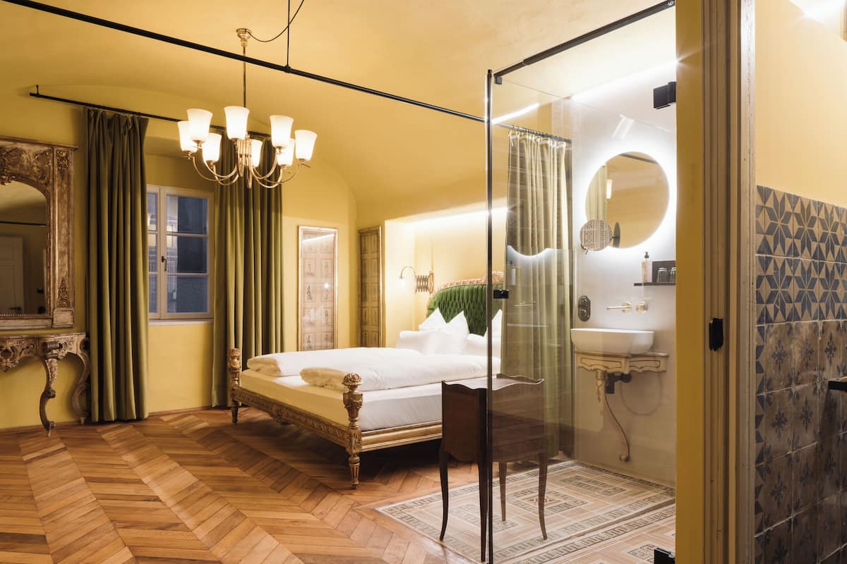 Ein Schlafzimmer mit einem großen Bett, einer Kristalllüster, einer Glasdusche und einem Badezimmer mit Waschbecken und Spiegel.