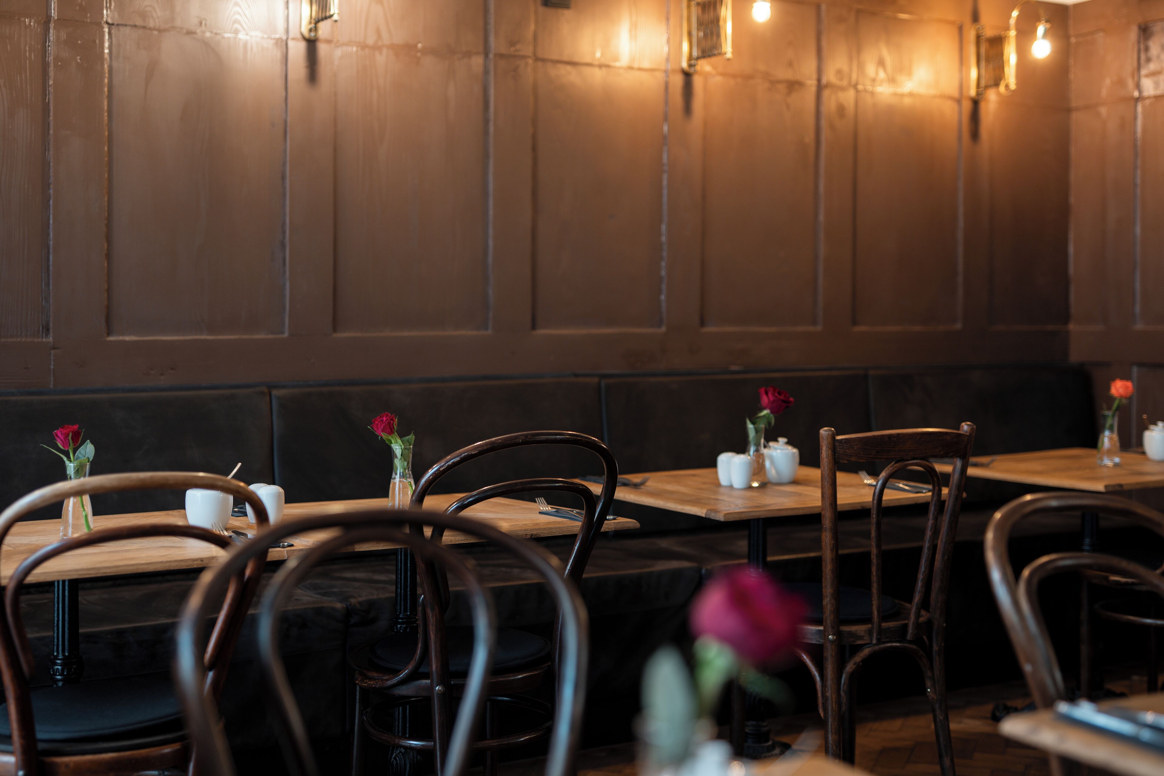 A restaurant interior with wooden tables and chairs, some with rose vases.