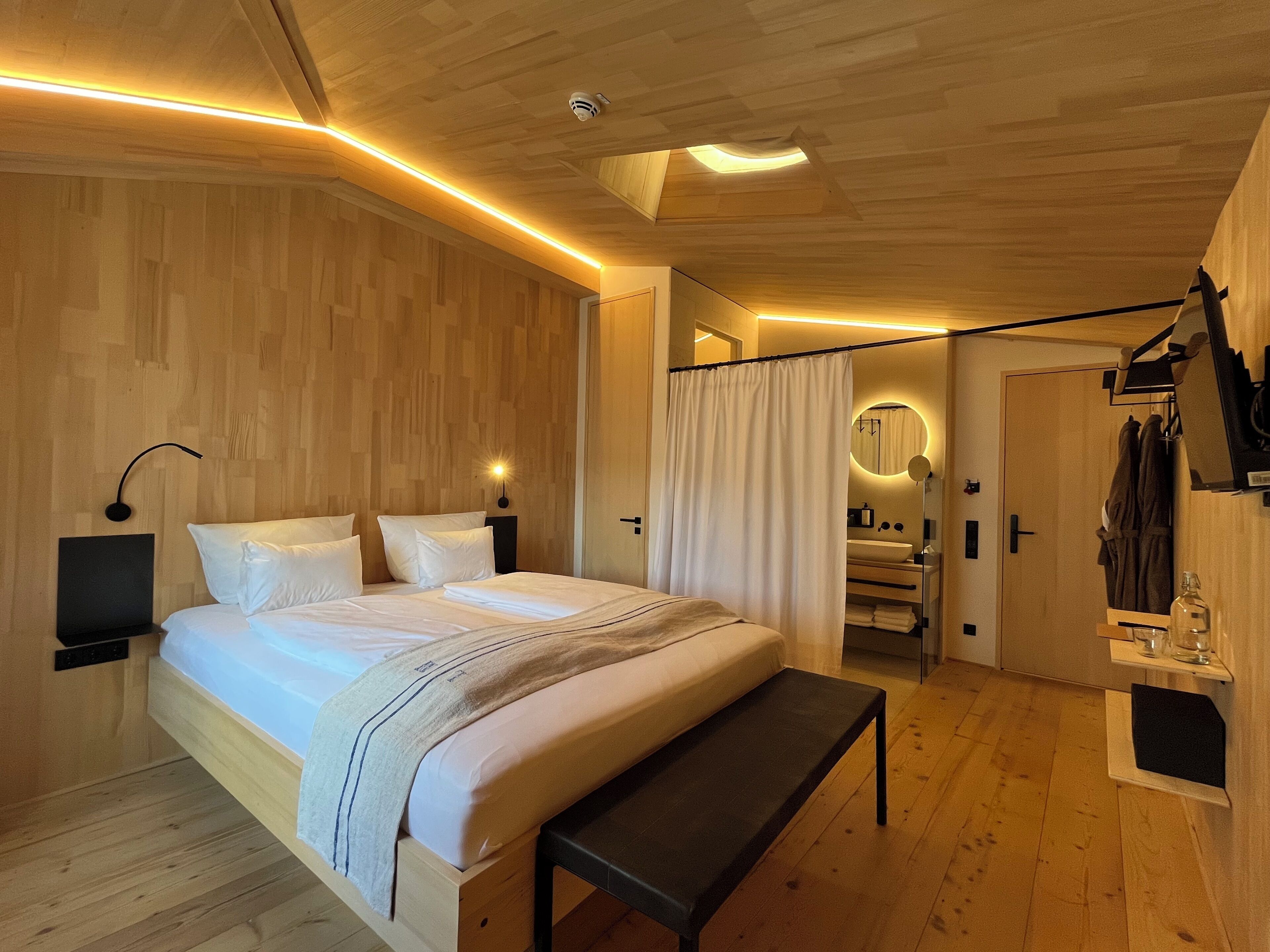 A modern bedroom with a wooden ceiling, a large bed with white bedding, a bedside table, a bench, and a bathroom visible through a curtain.