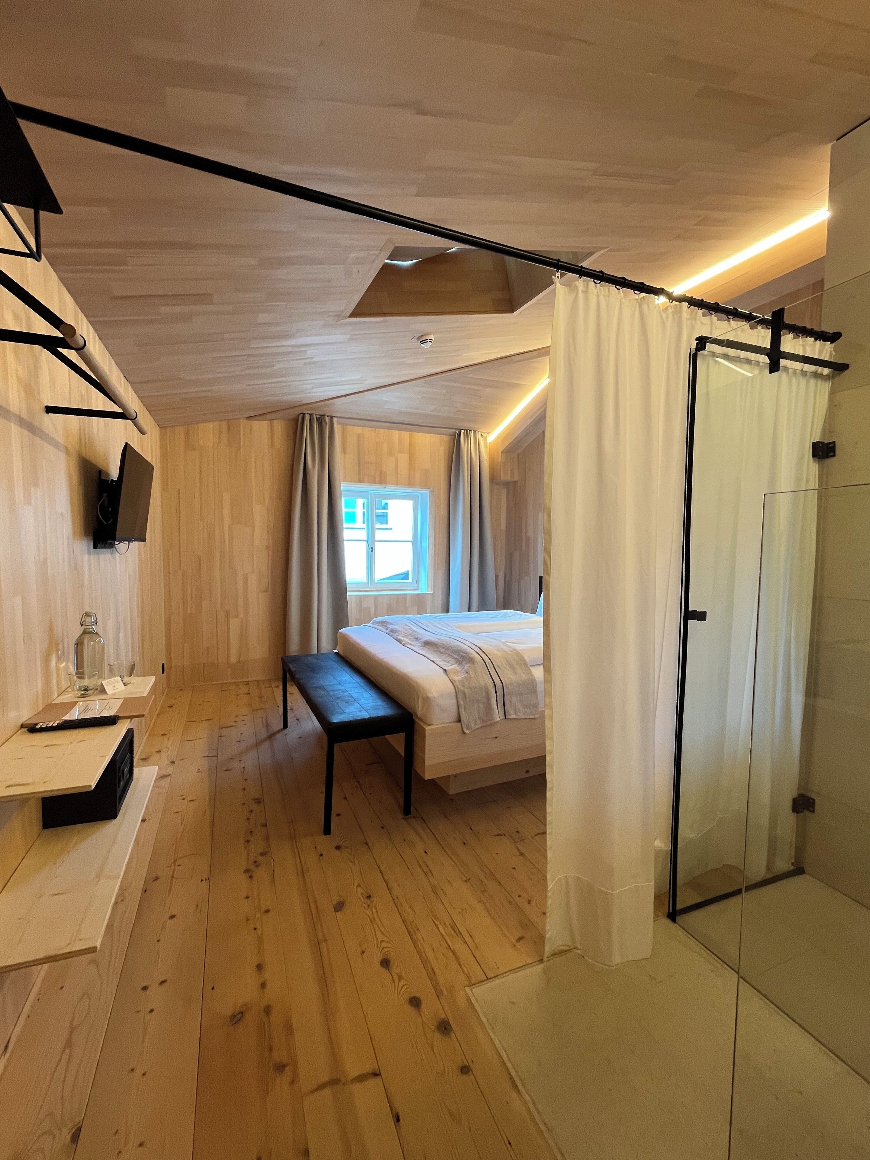 A bedroom with a wooden floor, a bed with a bench, a TV, a glass shower, and a window with curtains.