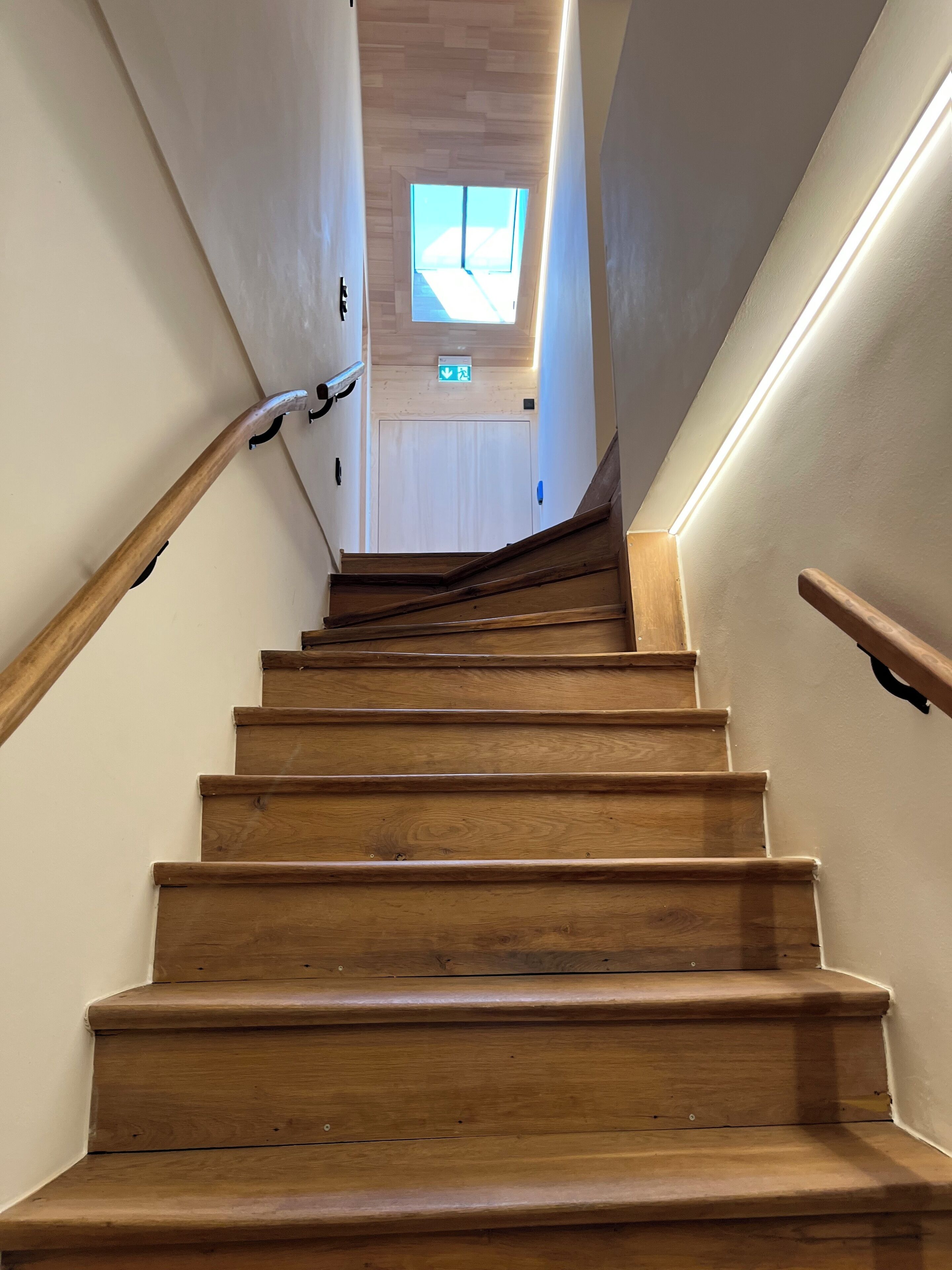 A wooden staircase with a handrail on both sides, leading to a door with a green exit sign.