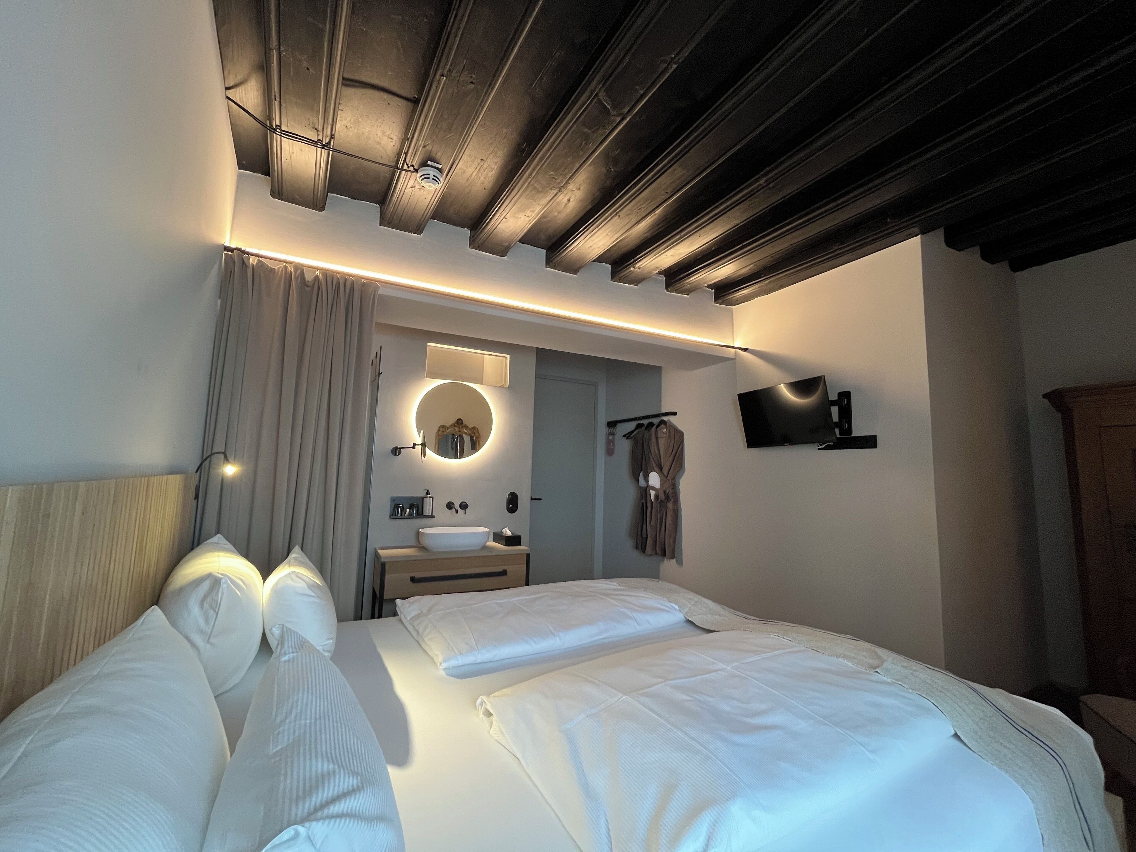 Quarto de hotel moderno com uma cama grande, um espelho redondo, uma televisão de tela plana e teto de madeira.
