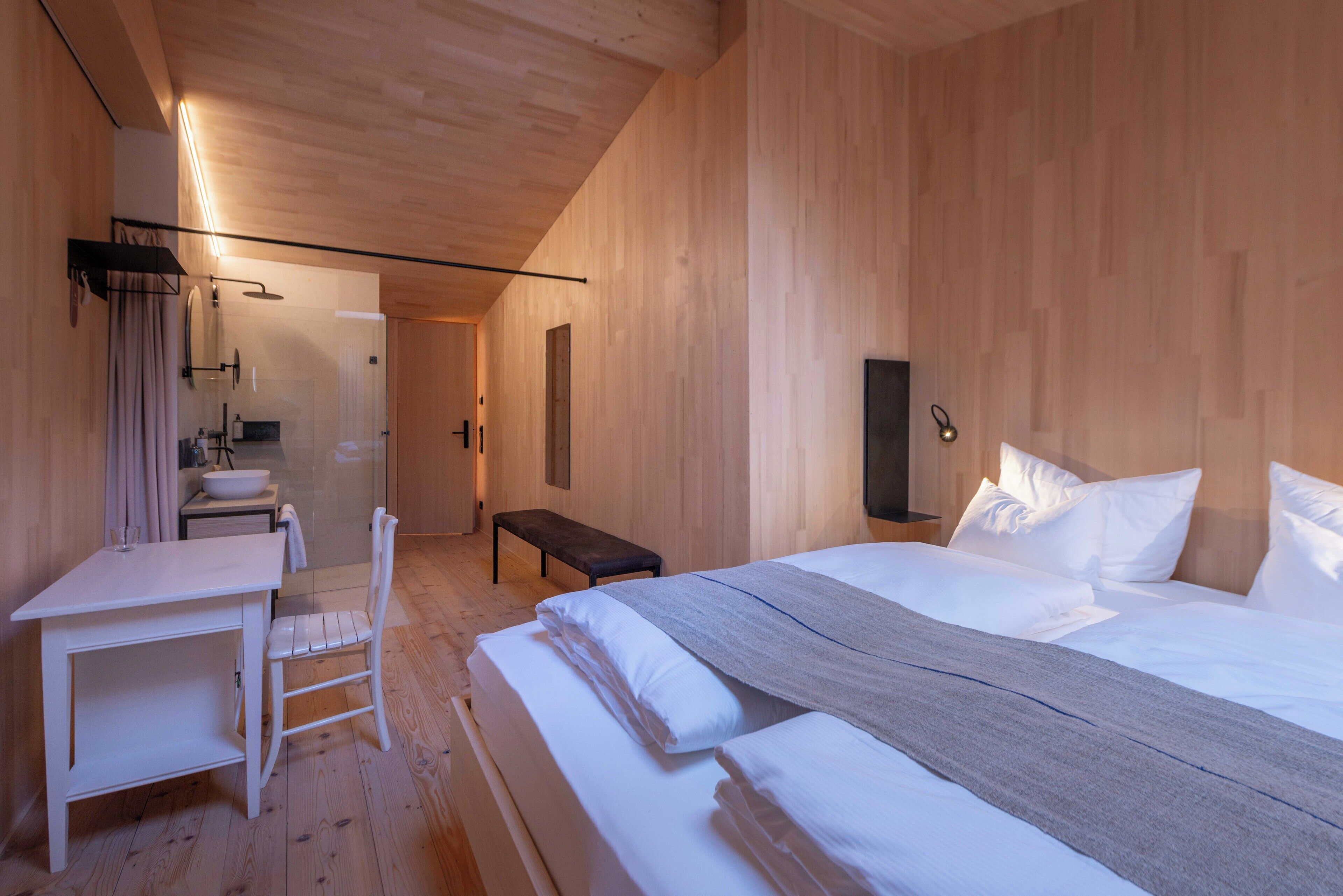Ein modernes Hotelzimmer mit Holzboden, einem Bett mit weißer Bettwäsche, einem Nachttisch, einem Sessel, einer kleinen Bank, einem Spiegel und einer Wandleuchte.