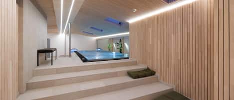 Un interior moderno con una escalera de madera que conduce a un área de spa, incluyendo una bañera de hidromasaje y un rincón de descanso con cojines.