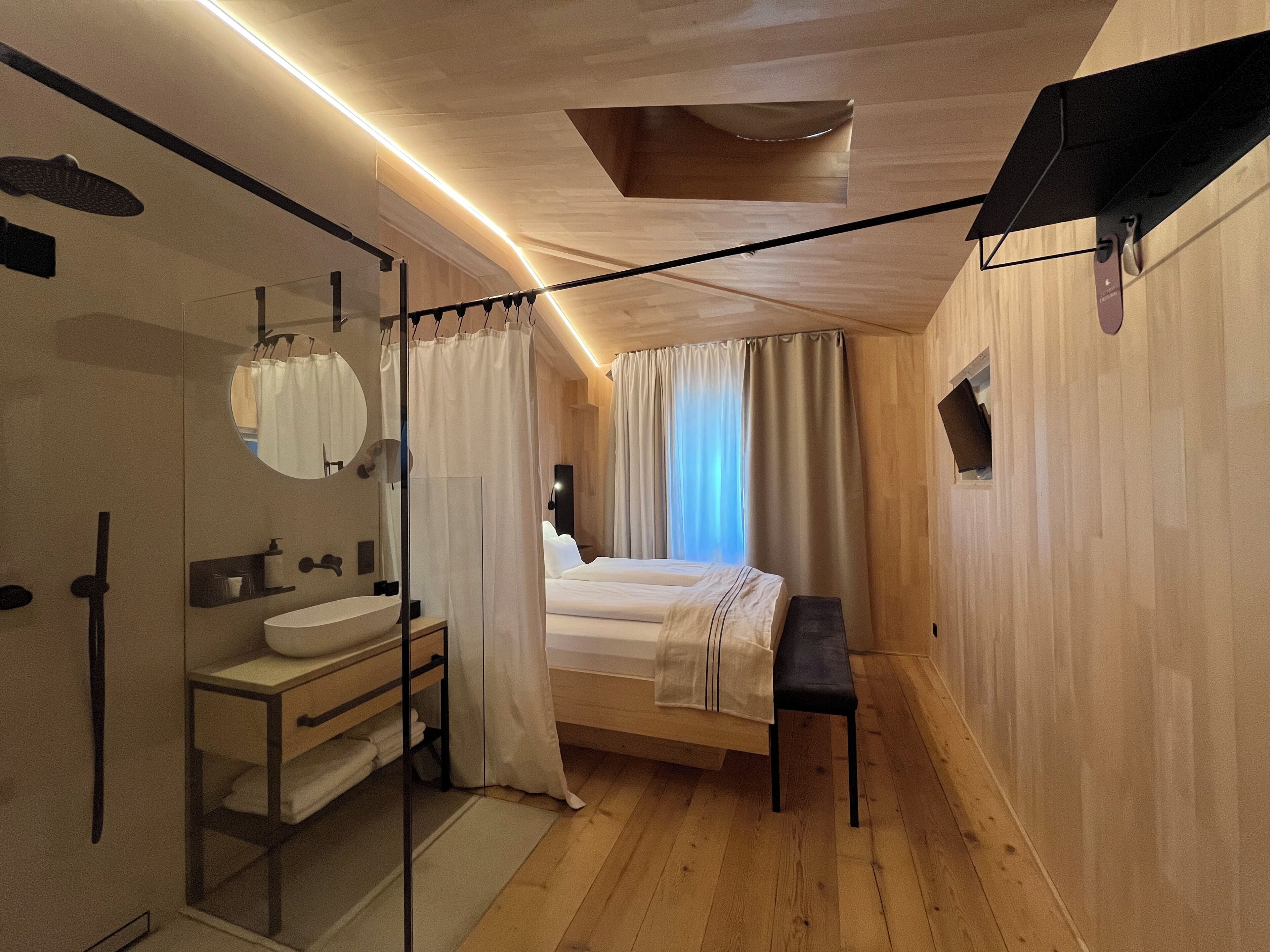 Un dormitorio moderno con techo de madera, una cama grande, una mesita de noche, un espejo y una ventana con cortinas.