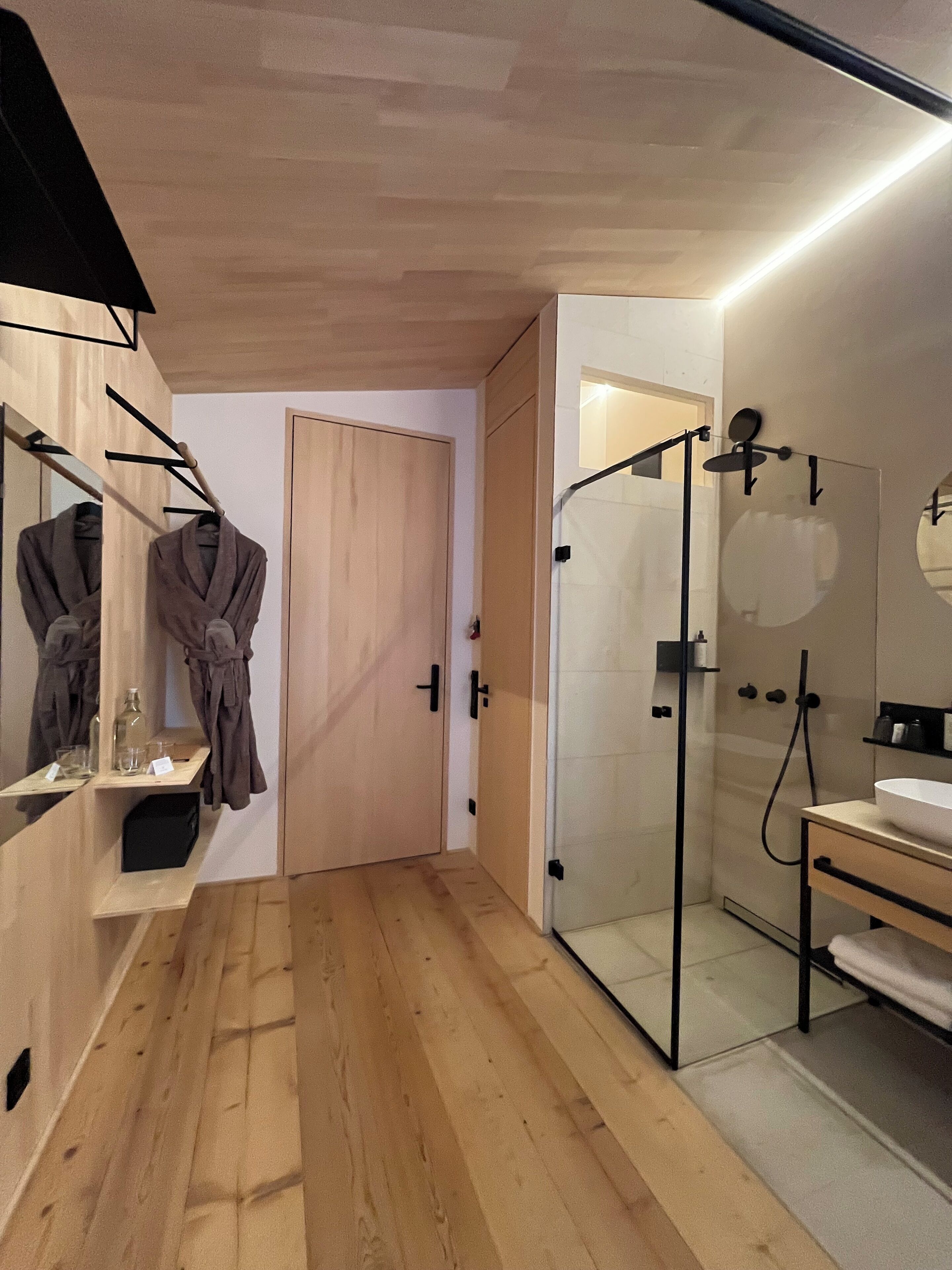 Une salle de bain moderne avec un plancher en bois, une douche en verre, un miroir avec un porte-peignoir et un lavabo avec un porte-serviettes.