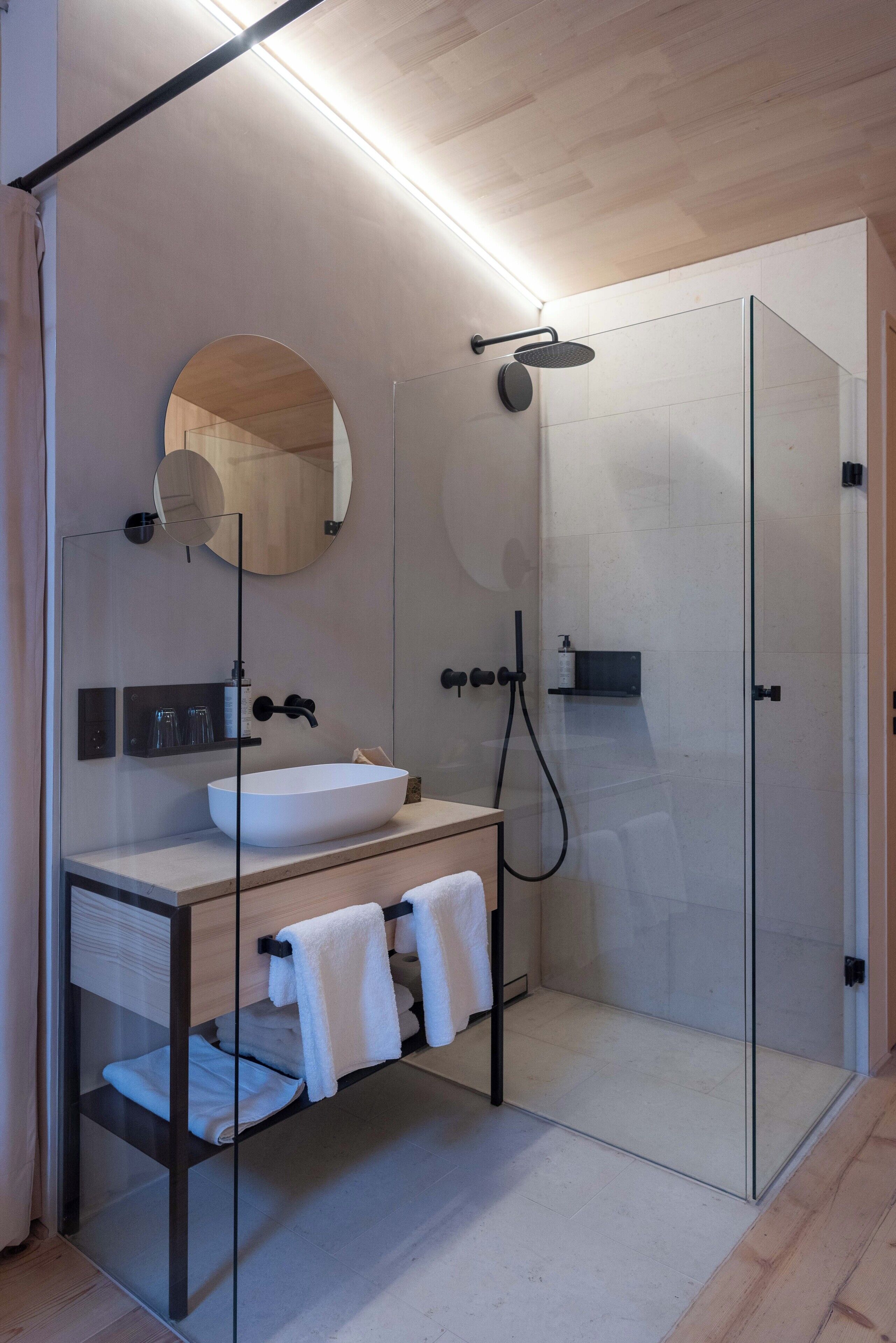 Un baño moderno con ducha envuelta en vidrio, un espejo redondo y un mueble de tocador de madera con lavabo blanco.