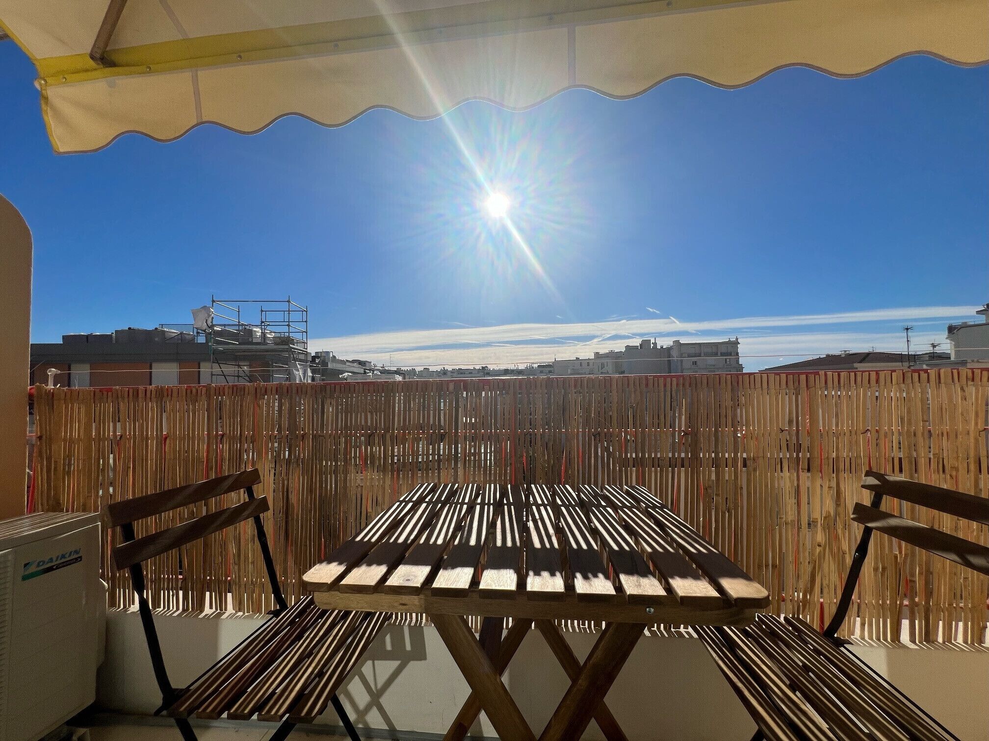 Een terras buiten met een houten tafel en stoelen, een plafondventilator en een uitzicht op gebouwen op de achtergrond.