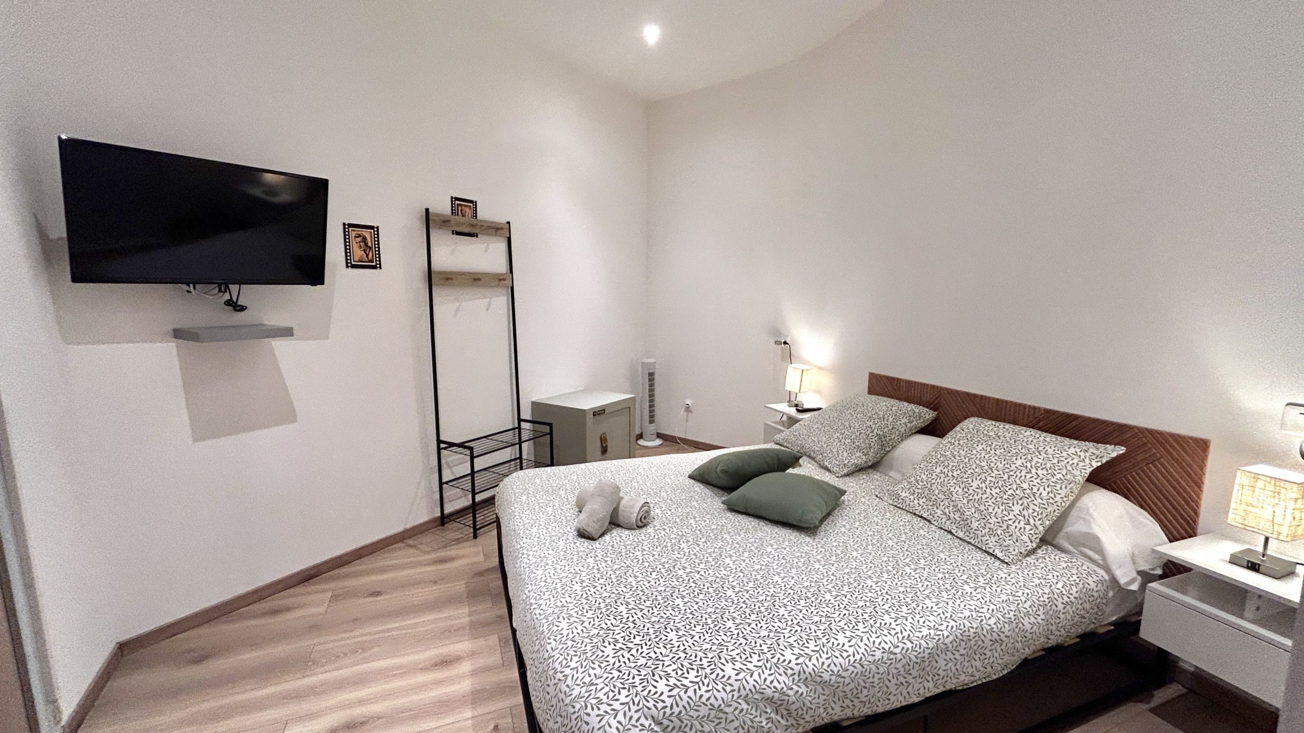 A neatly arranged hotel room with a bed, a TV mounted on the wall, a bedside table with a lamp, and a small bedside cabinet.