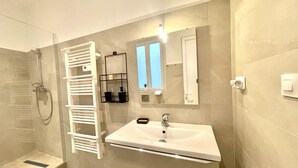 A modern bathroom with a glass shower enclosure, a white towel rack, a sink with a mirror above it, and a small window providing natural light. - woody (Cannes)