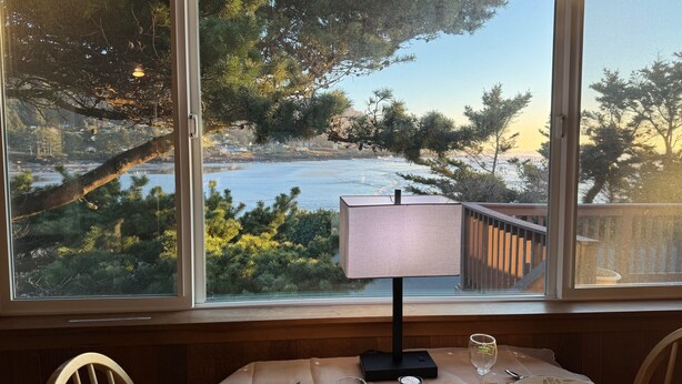 A table set with dinnerware and a wine glass, a lamp, and salt and pepper shakers, with a view of a coastal landscape through a large window.