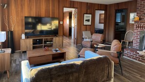 A living room with a large flat-screen TV, wooden panel walls, a brick fireplace, and a cozy seating area with a sofa and chairs.
