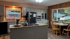 A modern kitchen with a central island, stainless steel appliances, and a view of a forest through a large window.