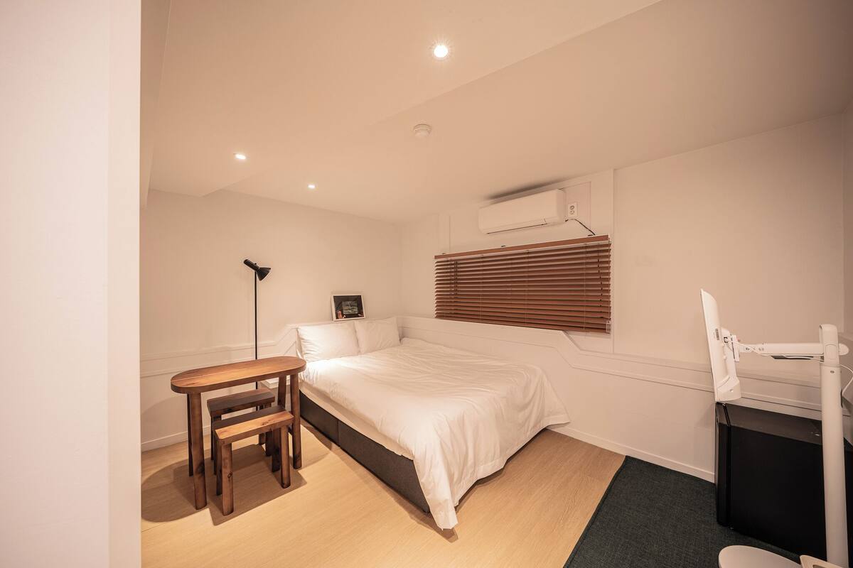 Ein Schlafzimmer mit einem Bett, einem Schreibtisch mit Stuhl, einer Stehlampe und einem Fenster mit Jalousien.