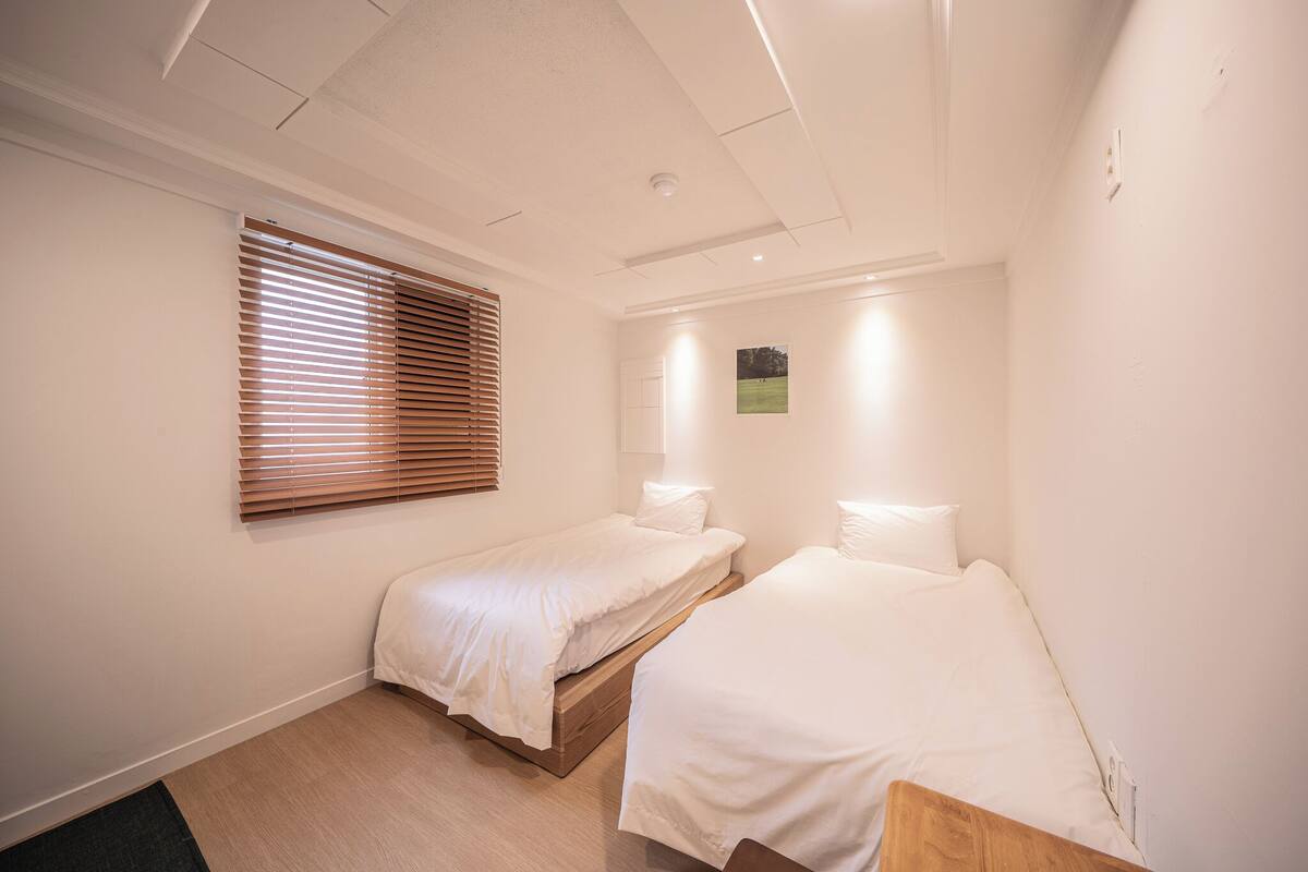 Ein kleines, sauberes Zimmer mit zwei Einzelbetten, einem Fenster mit Jalousien, einer kleinen Wandleuchte und einer Tür.
